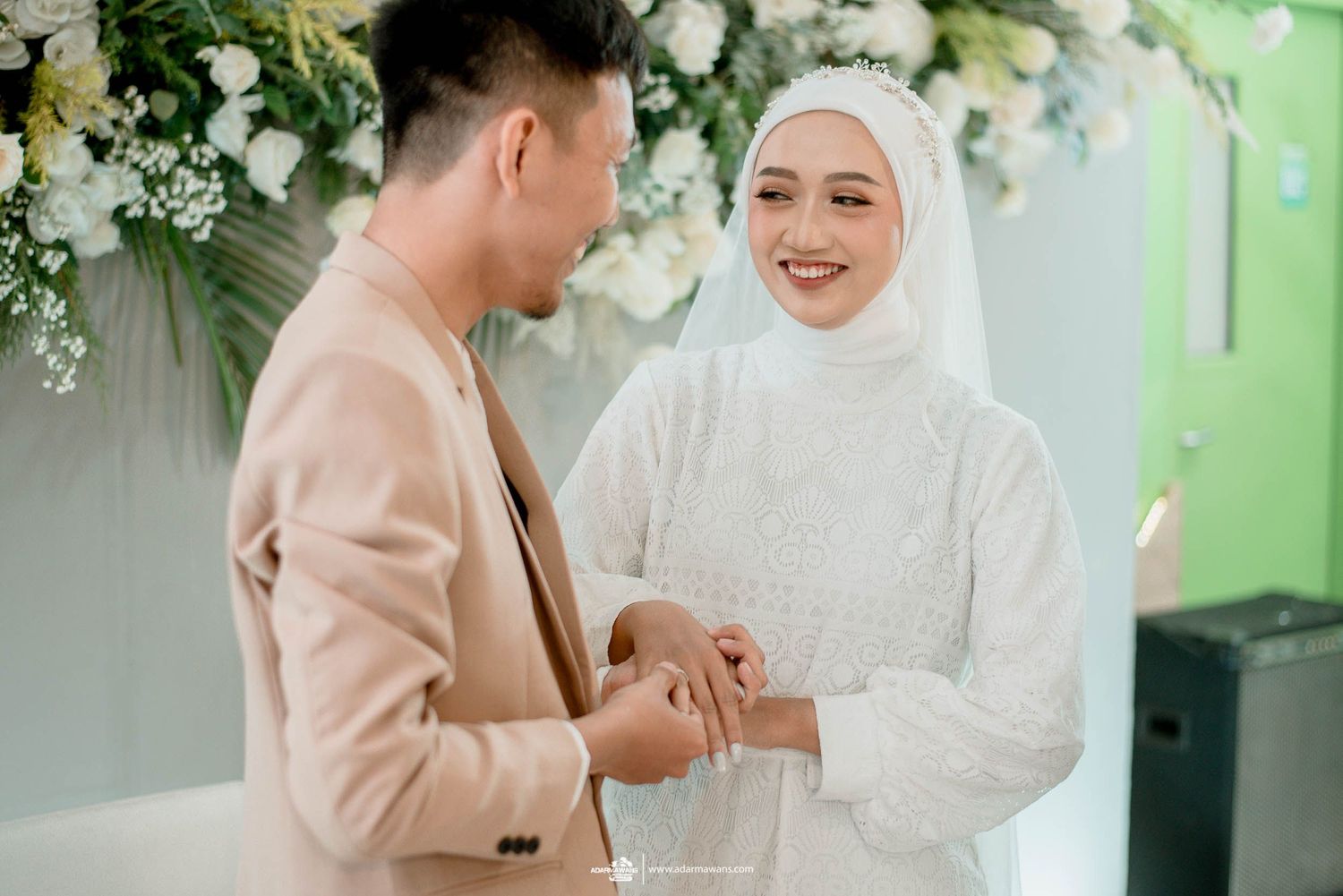 Bride and groom exchange rings during a wedding ceremony under white floral arch with green backdrop.