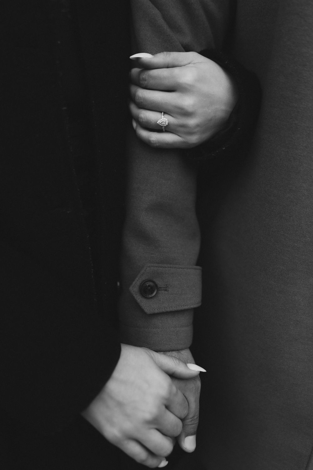 Black and white photo of hands with engagement ring against a coat sleeve.