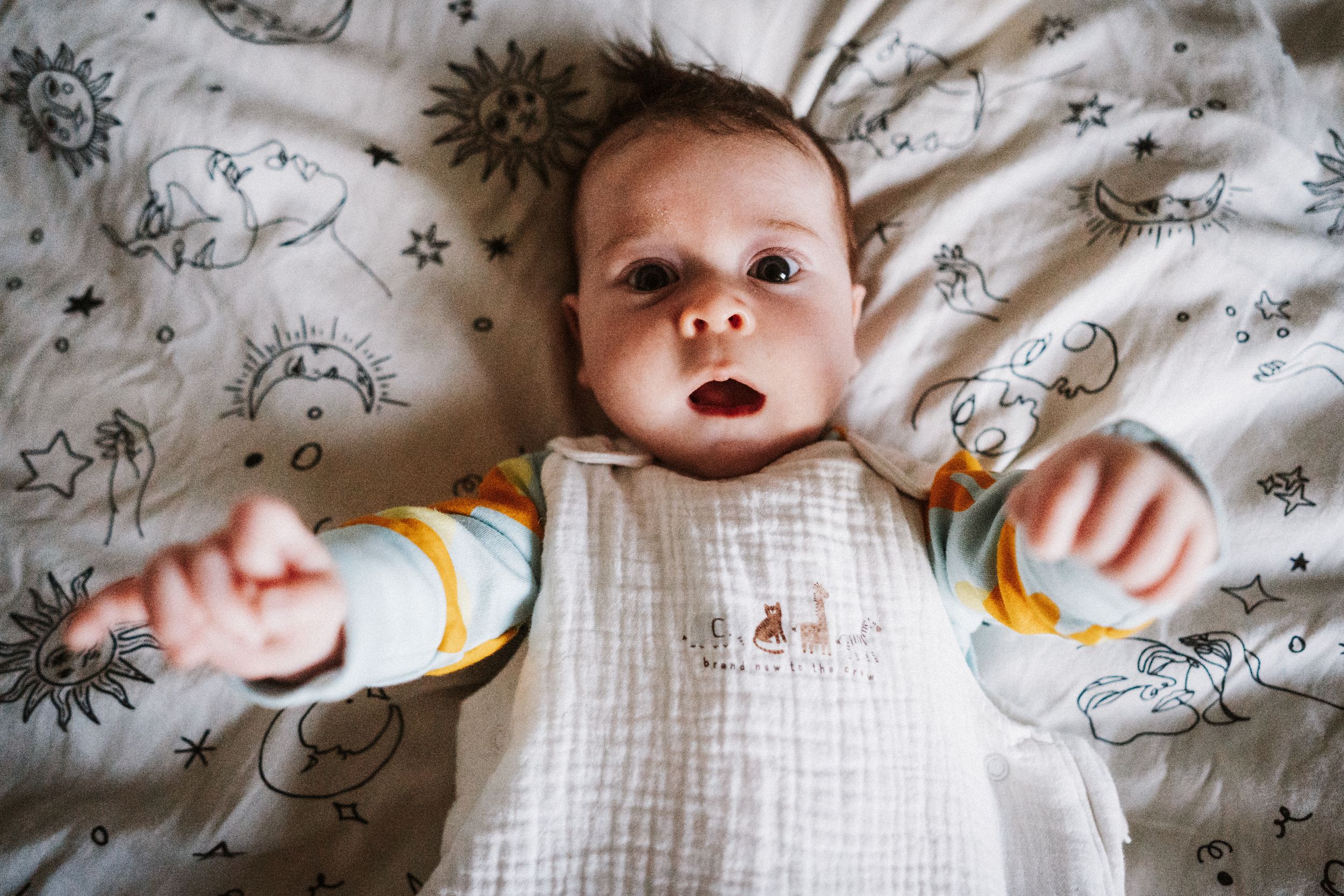 Baby wearing white onesie lies on floral patterned bedding while reaching out with small hands.