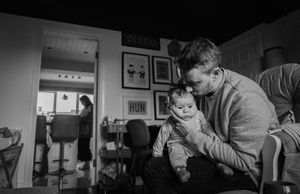 A black and white photograph shows a tender moment with a baby in a cozy home interior while someone works in background.