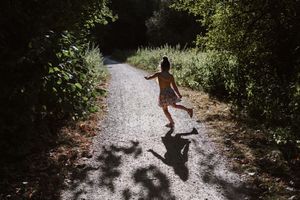 Person walking along a sunlit forest path casting long shadows through trees and dappled light.