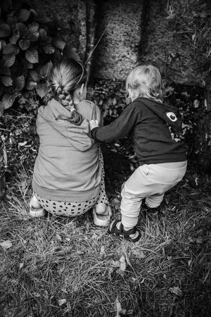Two small children sit together in black and white on grass near bushes in matching outfits.