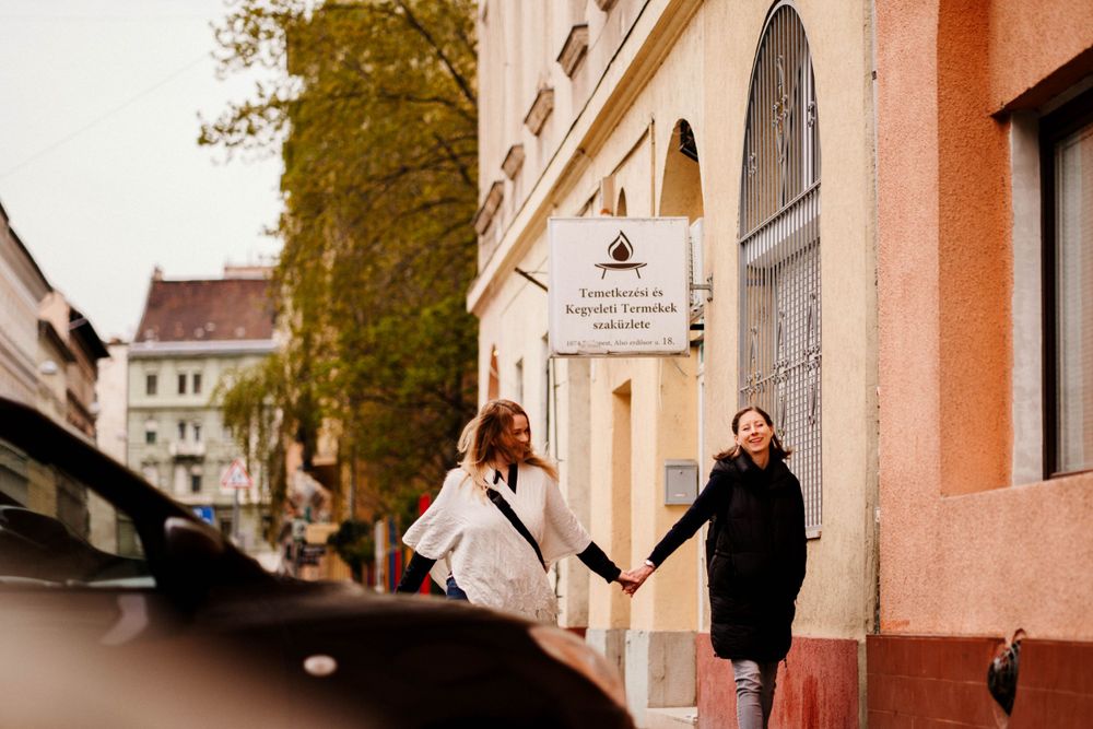 Two people holding hands outside a pink building with arched windows on a tree-lined street.