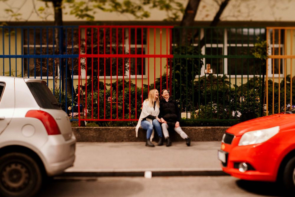 A couple sits between parked cars on a city street with red and silver vehicles visible.