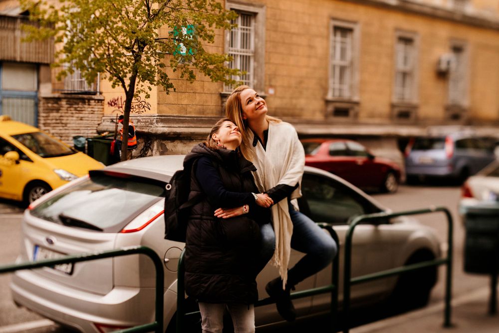 Two people laughing while leaning against metal railings with parked cars in background.