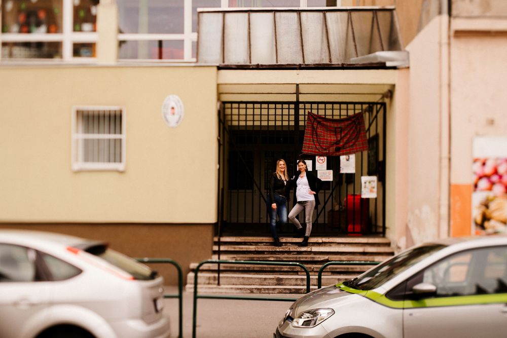 Two people standing in a building entrance between parked cars on a residential street.