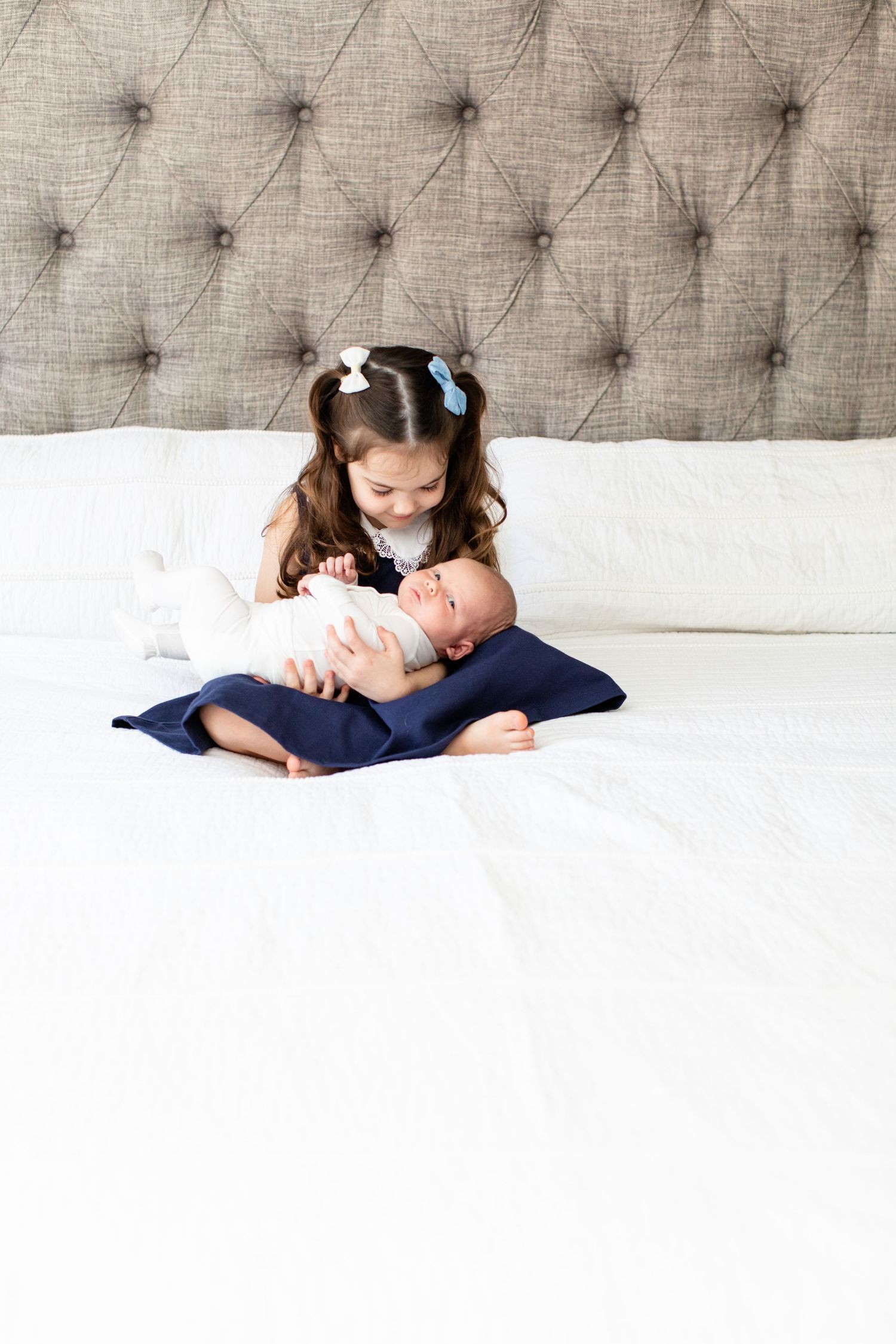 Young girl in white dress with blue bows holds newborn sibling on white bed.