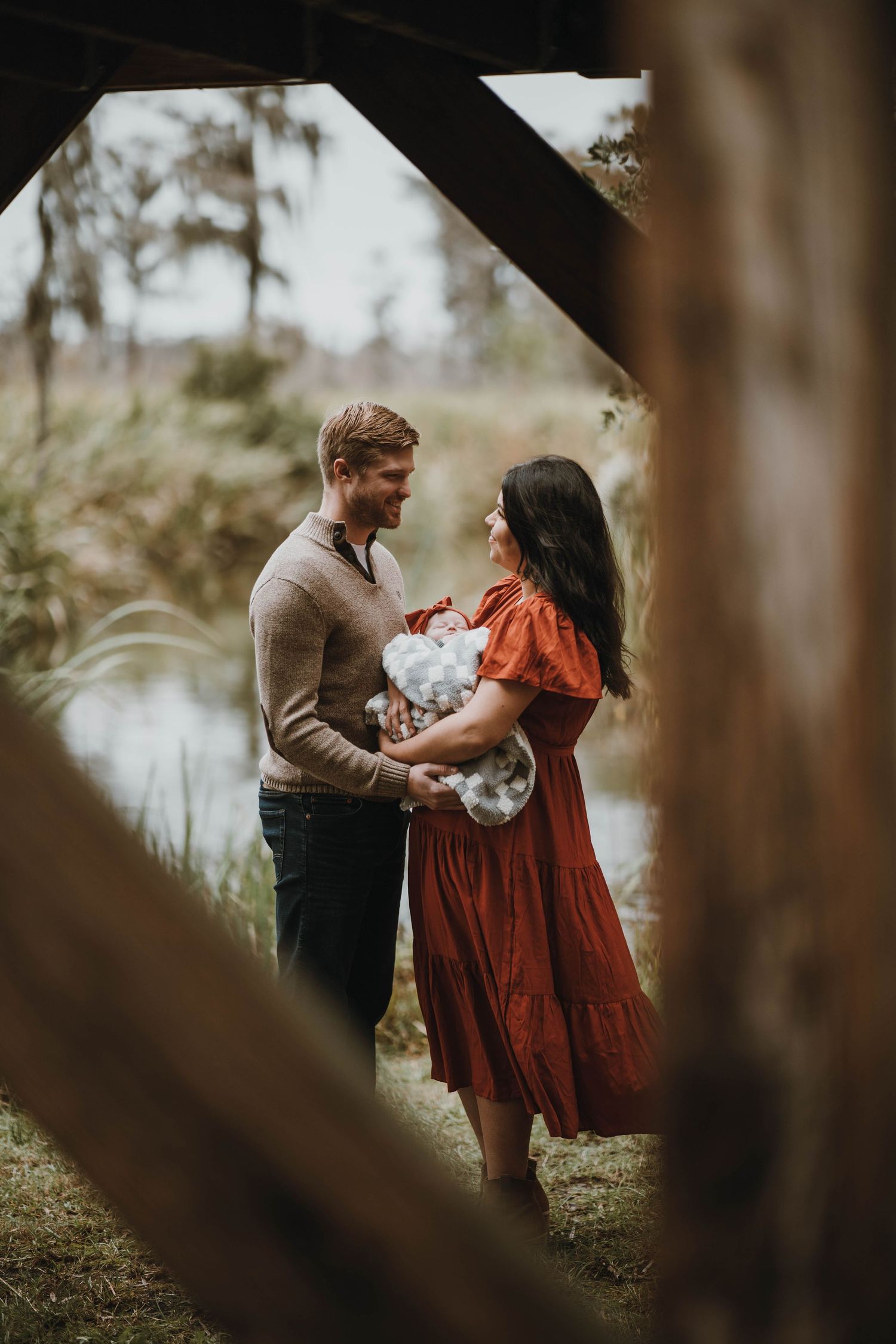 A romantic couple embraces outdoors while holding a bouquet of white flowers, framed through trees in soft lighting.