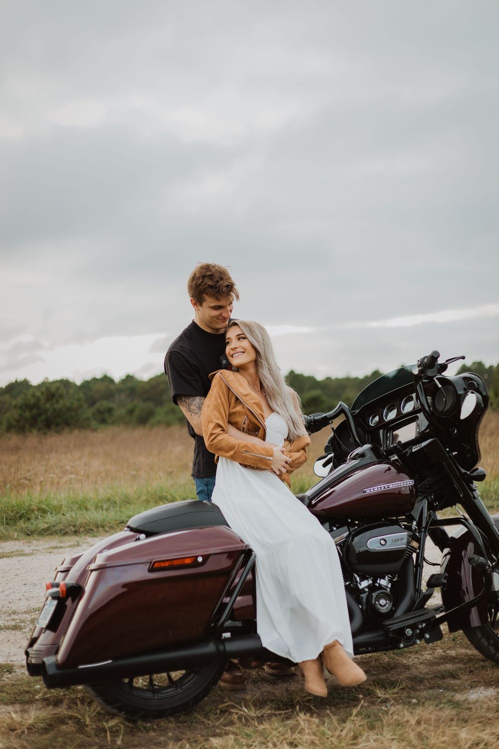 A couple poses together on a maroon Harley Davidson motorcycle in a rural field setting.