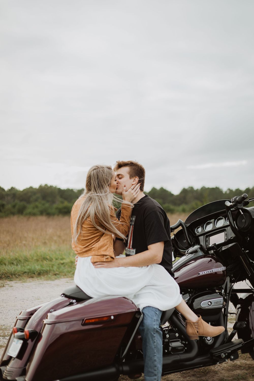 An intimate motorcycle photoshoot featuring a couple on a burgundy Harley against an overcast sky.