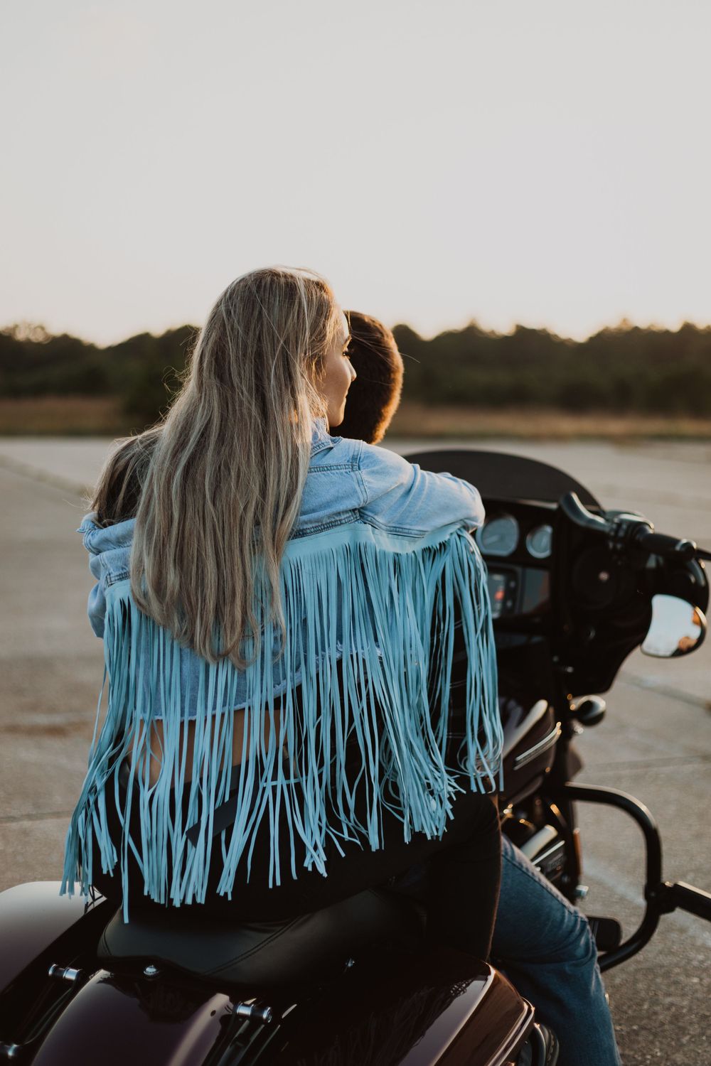 A person wearing a blue fringed jacket rides on a motorcycle during sunset.