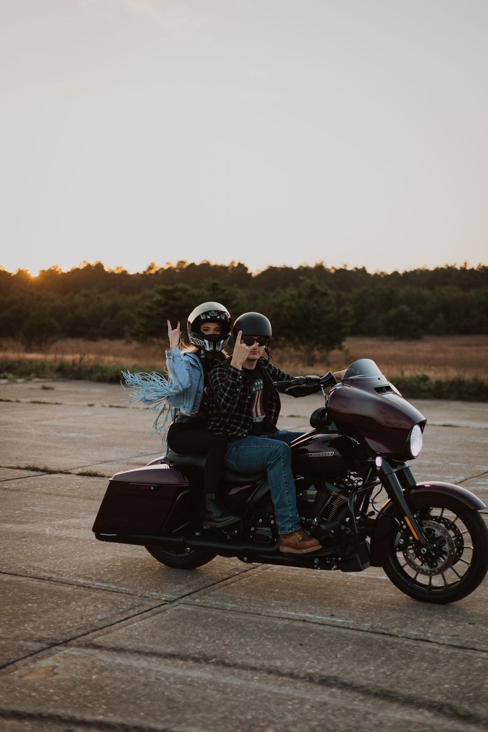 Two riders pose on a maroon Harley Davidson motorcycle during golden hour.