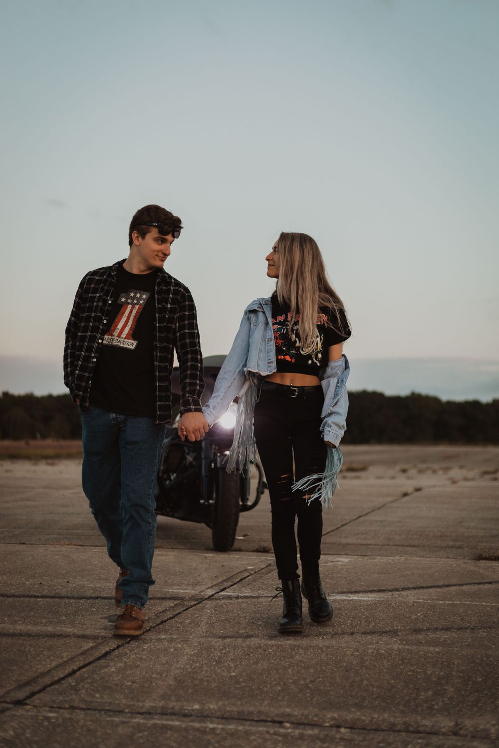 A casual couple stands beside a motorcycle on a concrete surface at dusk.