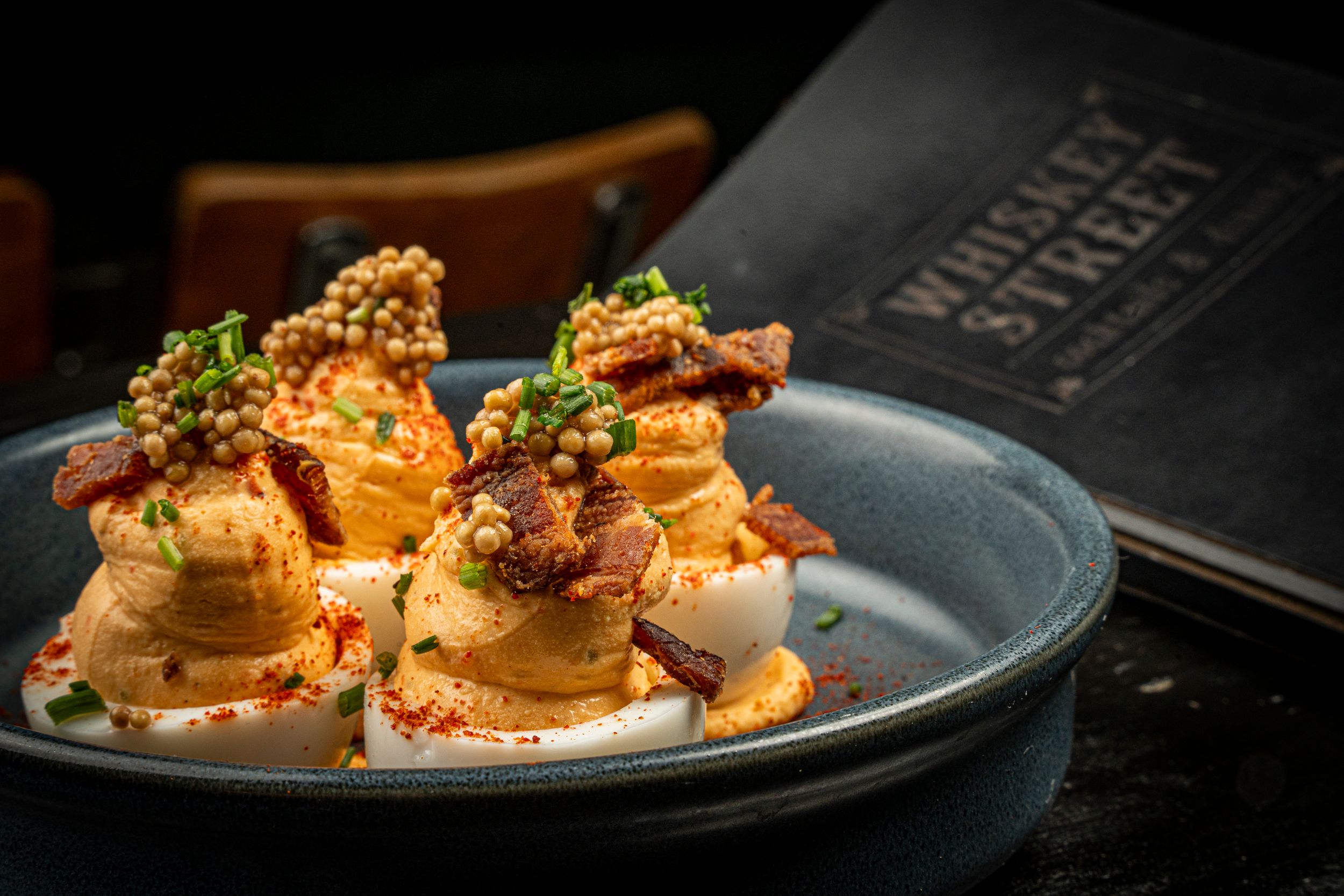 Elegant deviled egg appetizers garnished with crispy bacon and caviar pearls on blue plate at Whiskey Street.