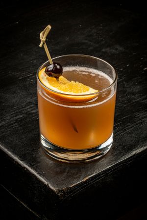 Amber cocktail garnished with an orange slice and dark cherry on a black wooden surface.