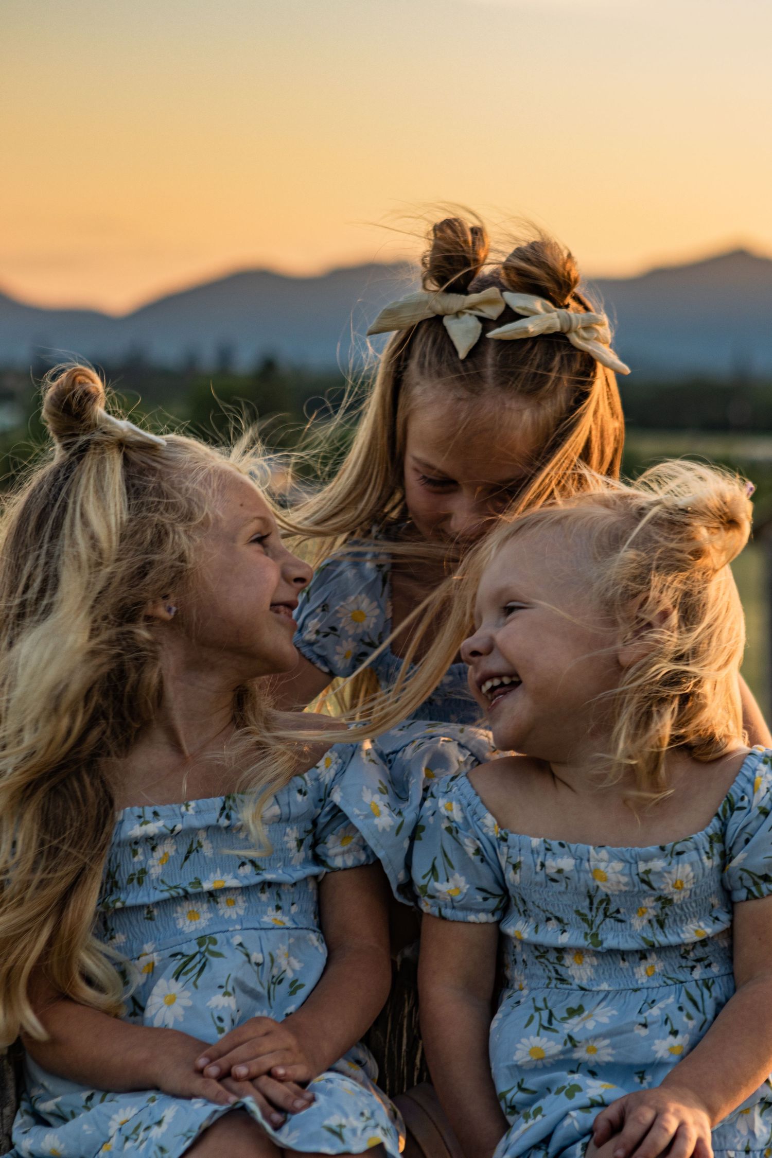 Three young blond sisters look at each other and giggle in the field.