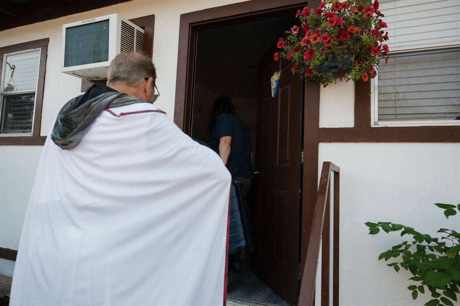 Someone wearing a white cape approaches a doorway decorated with red flowers.