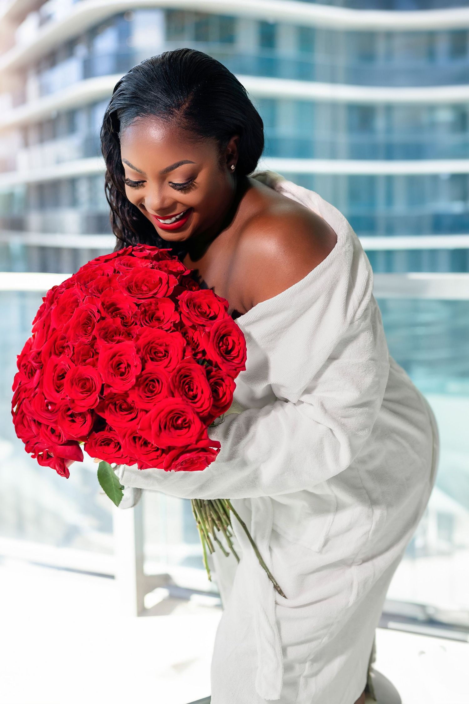 Smiling woman in white off-shoulder outfit holding large bouquet of red roses on balcony.