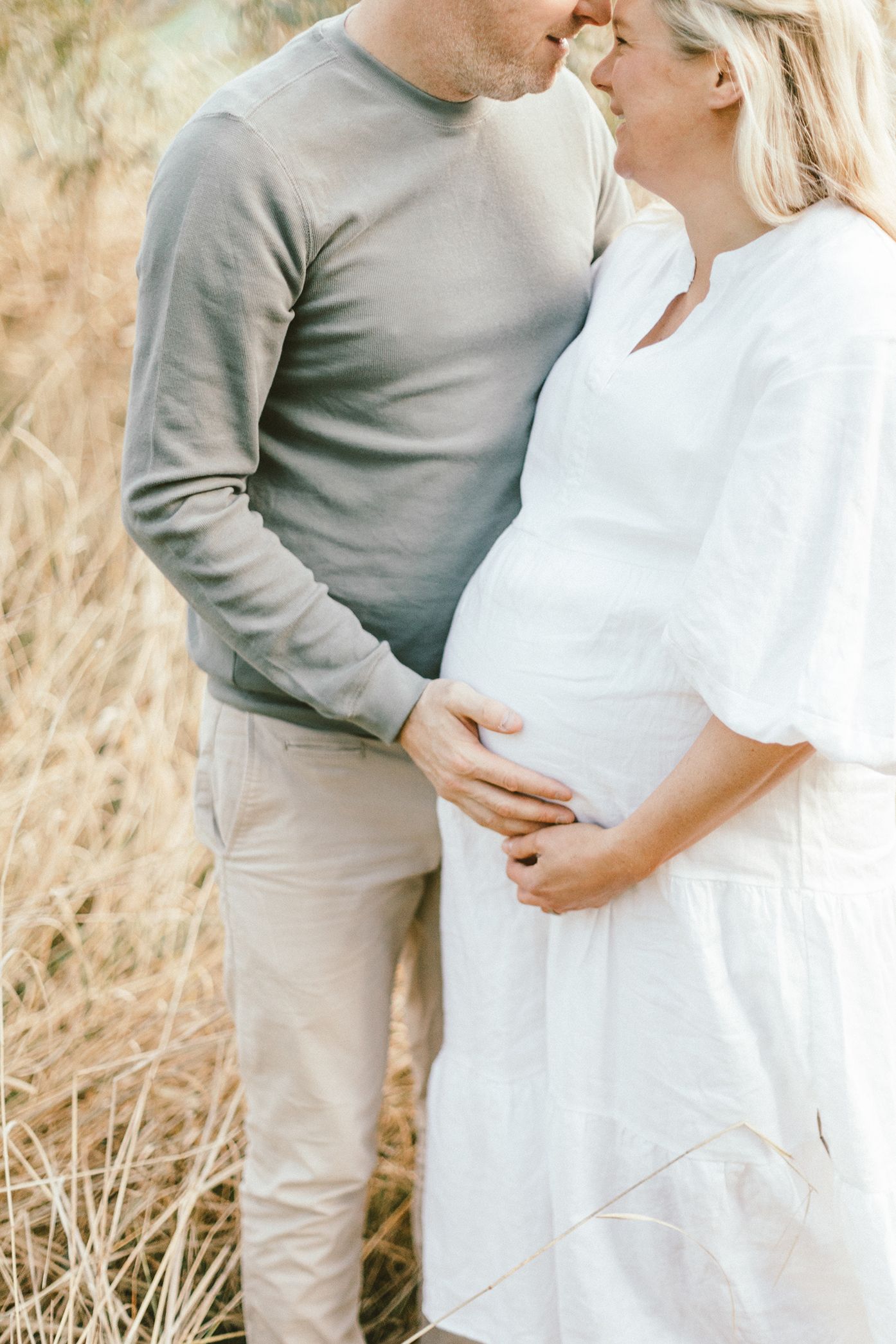 Intimate maternity photo of couple in white and gray clothing against natural backdrop.