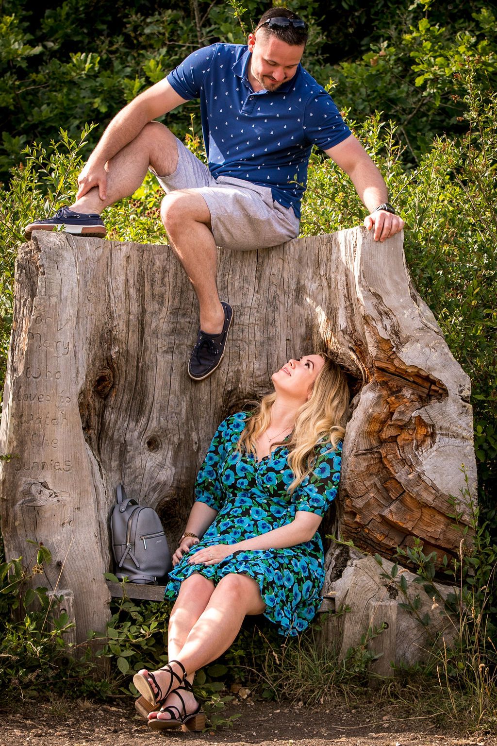 A playful couple posing on a large tree stump in a sunny outdoor setting.
