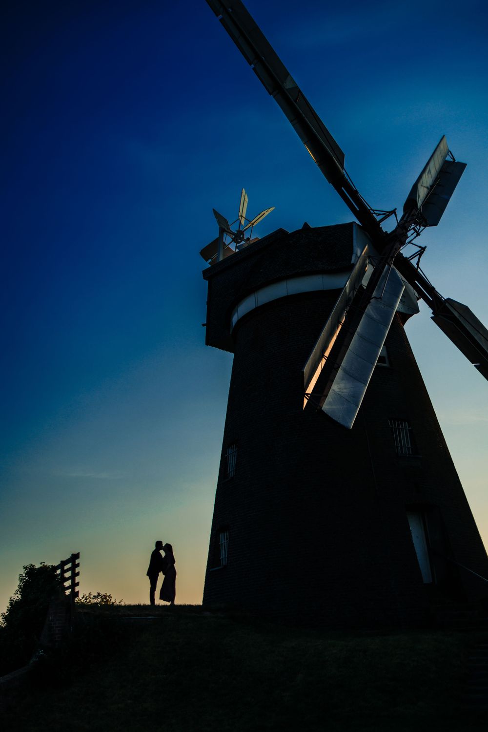 Silhouette of two figures standing by a traditional windmill against a dark blue sky at dusk.