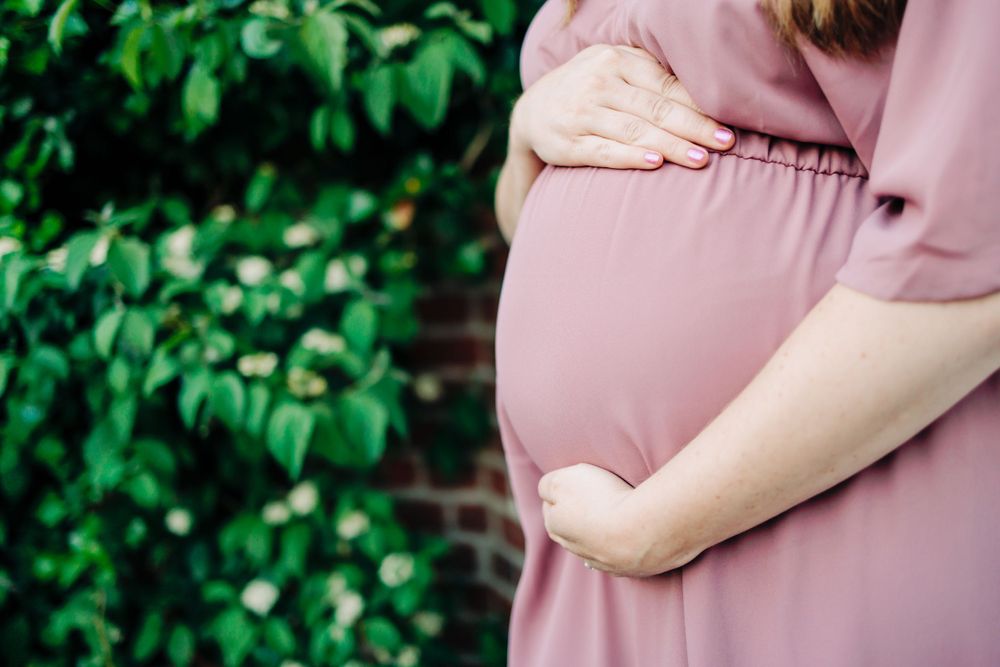Close-up of pregnant belly in flowing mauve dress against green foliage.