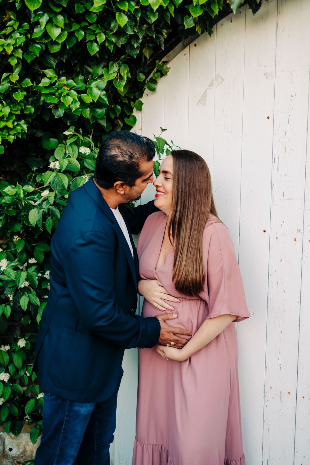Couple in formal wear embracing against white wall with green ivy.