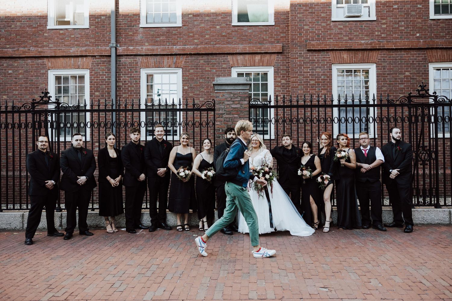 Bride and groom kiss while wedding party in black stands before iron fence on brick street. Cambridge, Ma Miranda Photo Co
