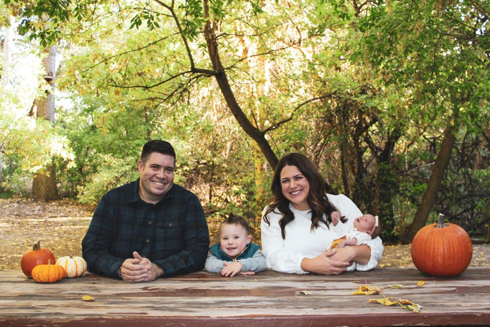 Family sitting at wooden table with pumpkins during fall photo session outdoors.
