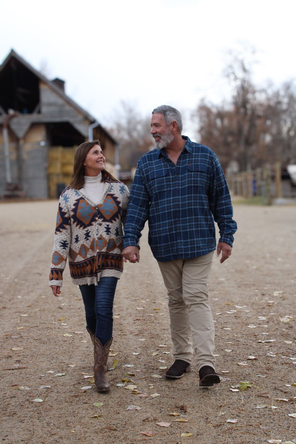 Couple walking hand in hand on dirt path near rustic building in autumn setting.