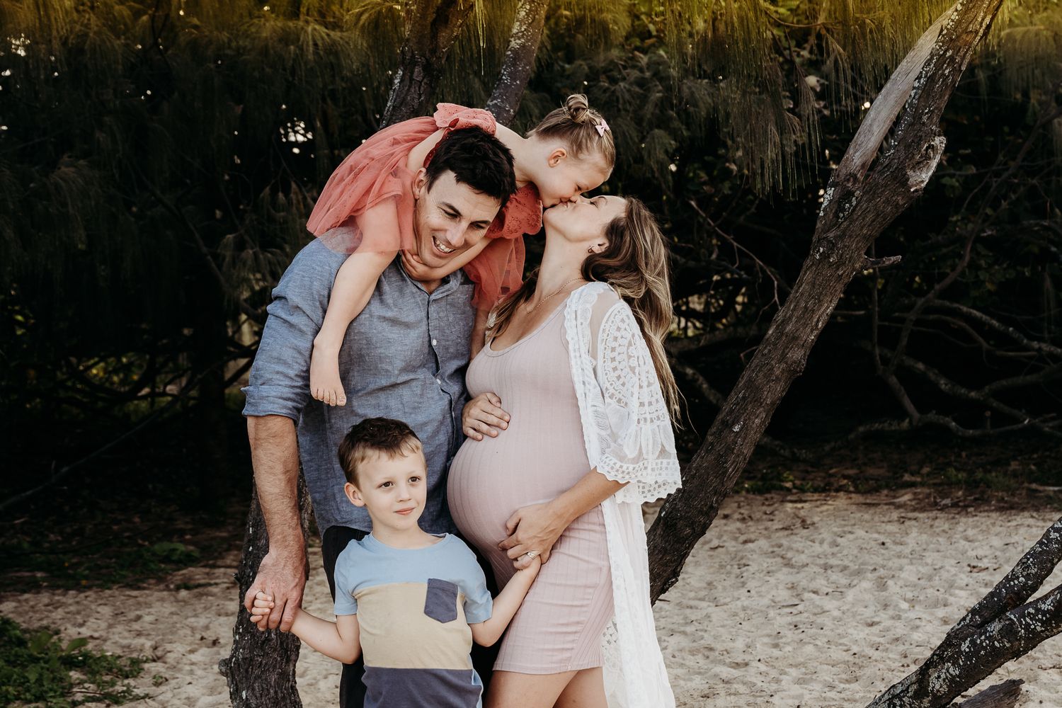 A tender family moment captured outdoors with an expecting mother in a white dress.