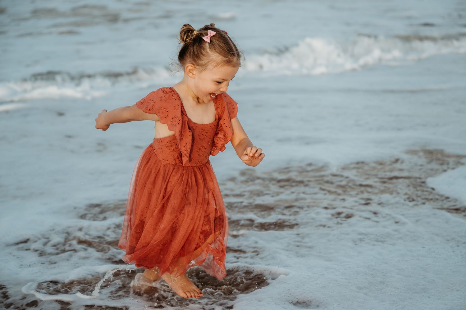 Little girl in coral dress dances joyfully in ocean waves.