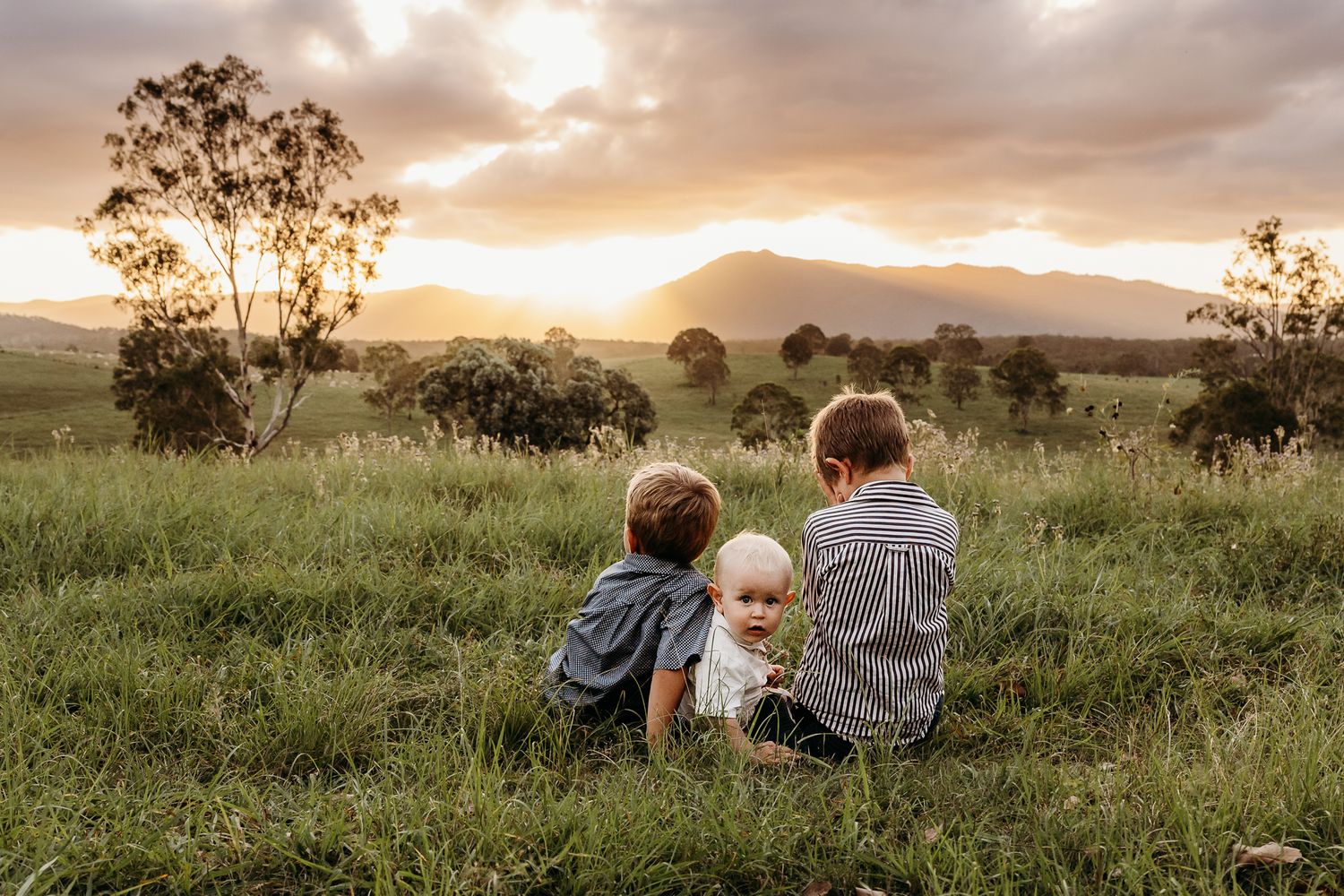 Three people sitting together on grass watching a beautiful sunset over mountain landscape.