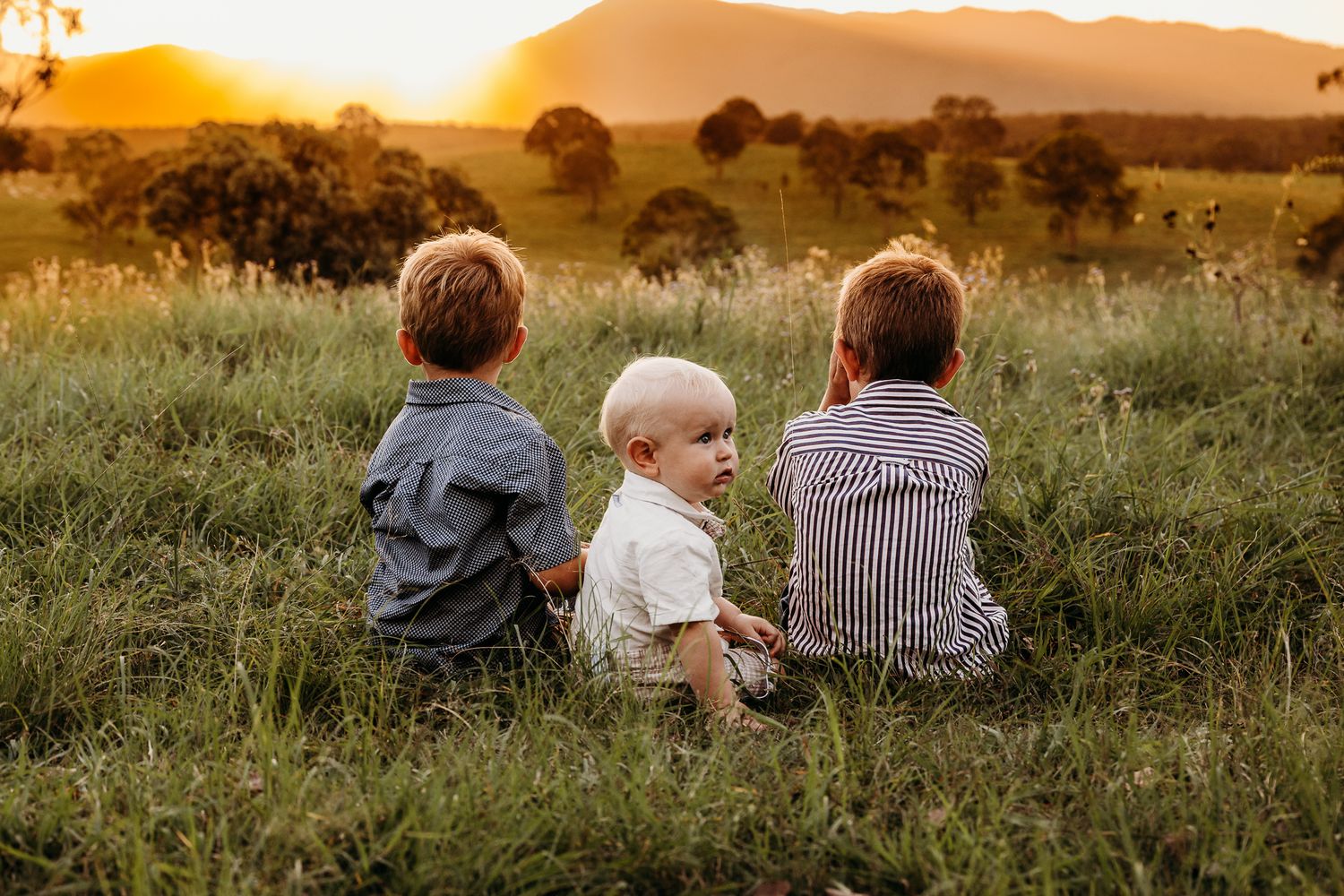 Three children sit together watching a beautiful golden sunset over rolling hills.
