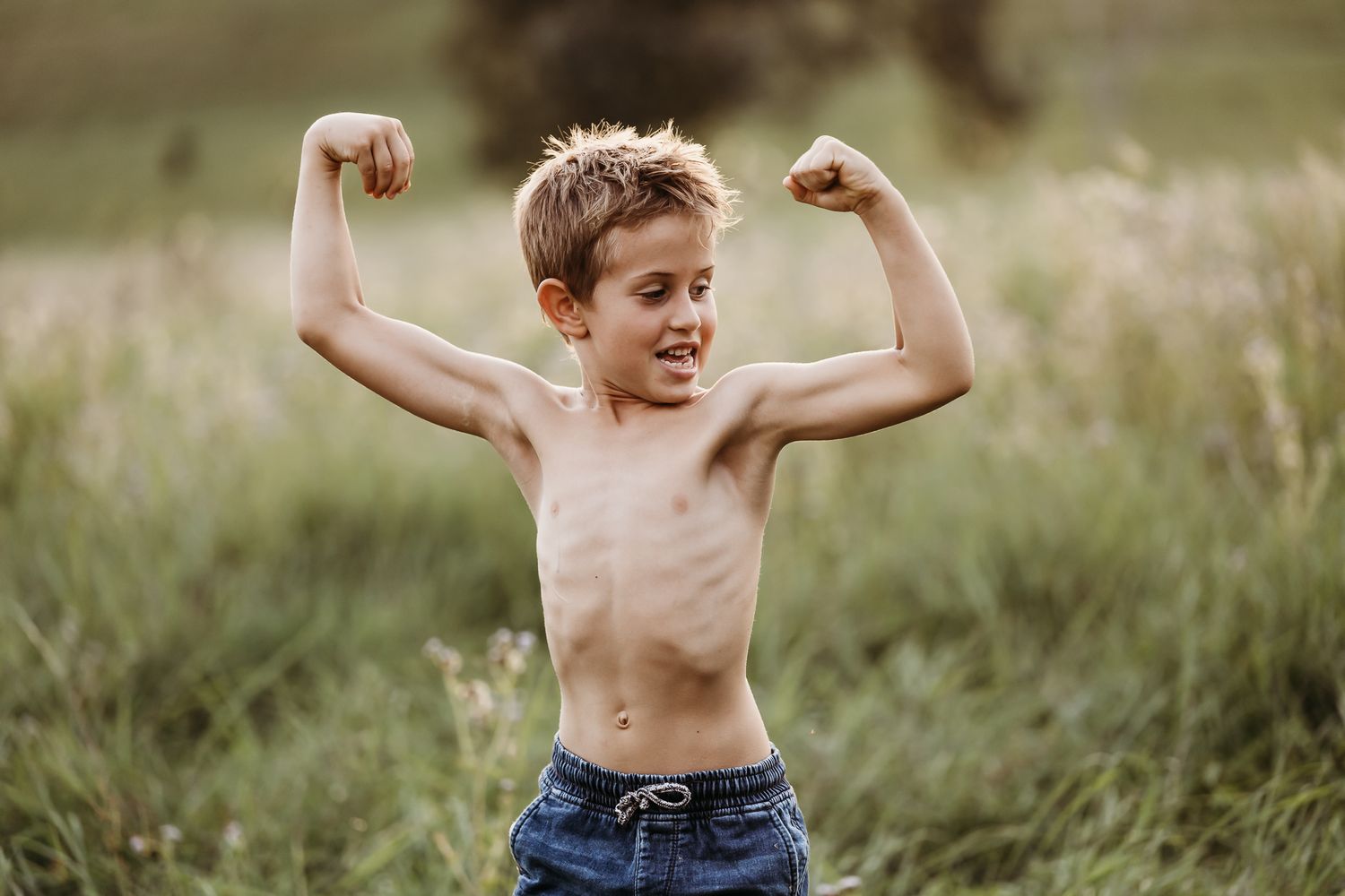 Young person flexing muscles while standing in tall grass at sunset.