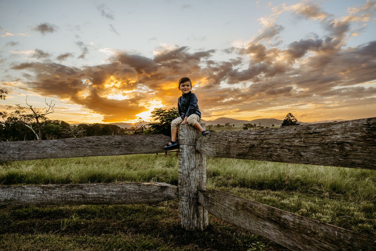 A child sits atop a wooden fence post during a dramatic golden sunset.