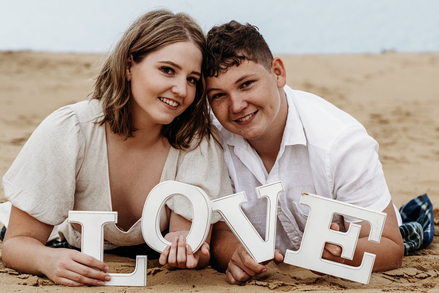 A couple lying on sand with white LOVE letters for an engagement photo shoot.