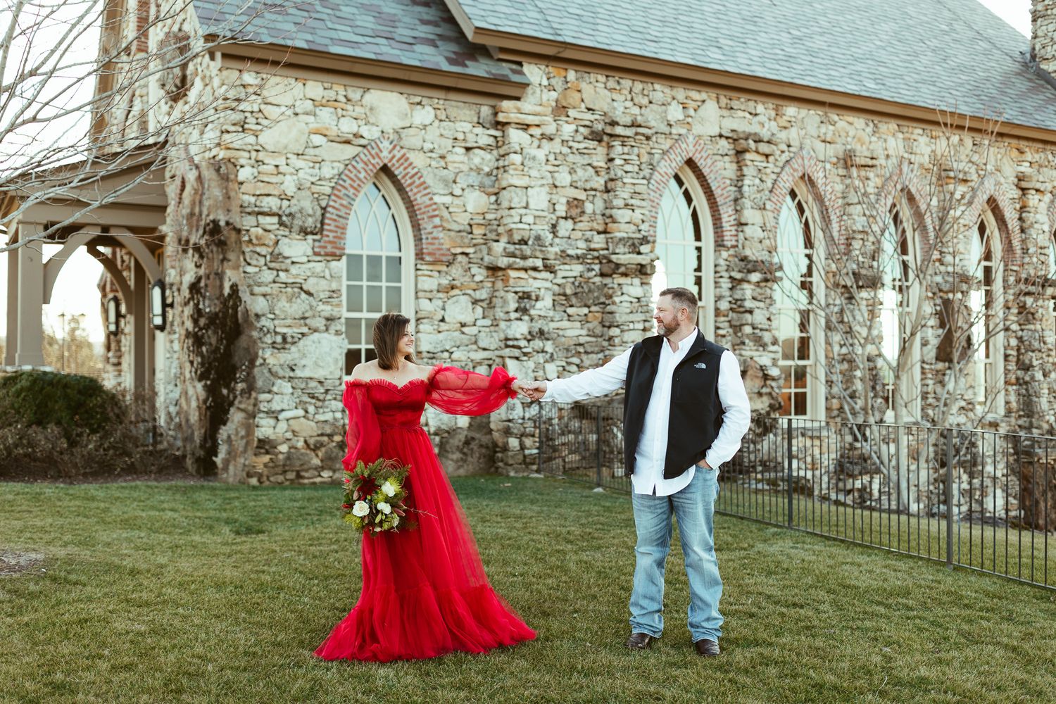 A couple poses in front of a stone church with arched windows, the woman wearing a flowing red dress.
