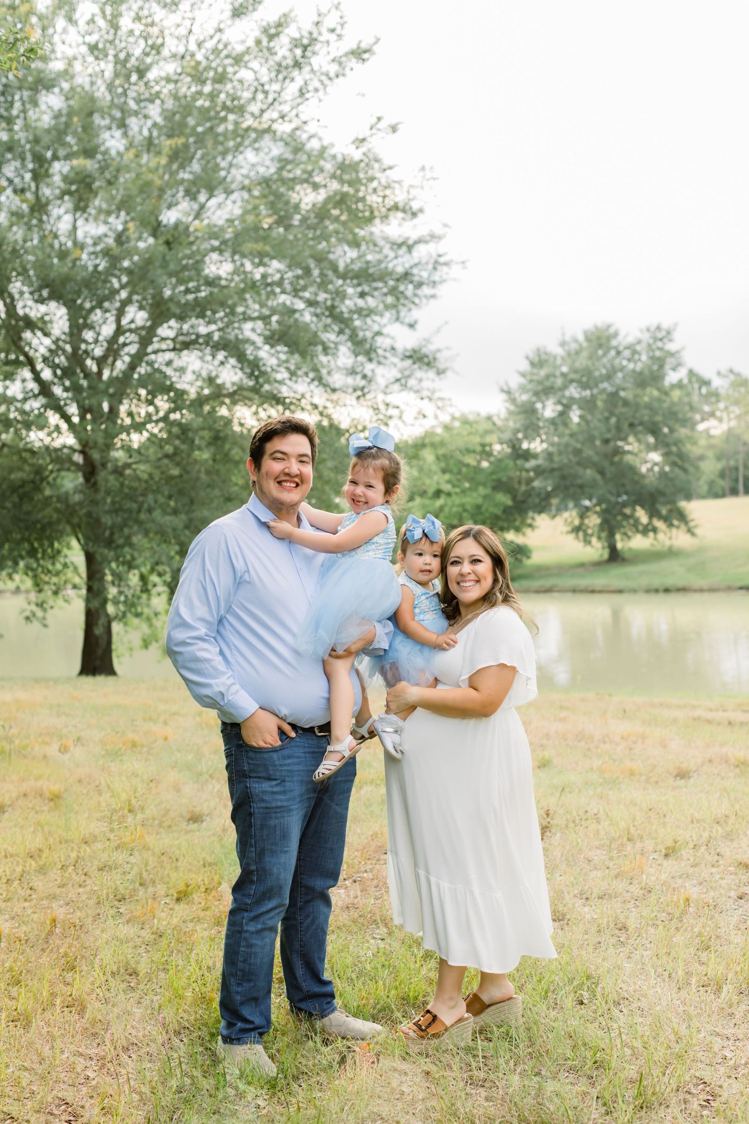 A family portrait in coordinated outfits standing together on grassy terrain with trees and a pond backdrop.