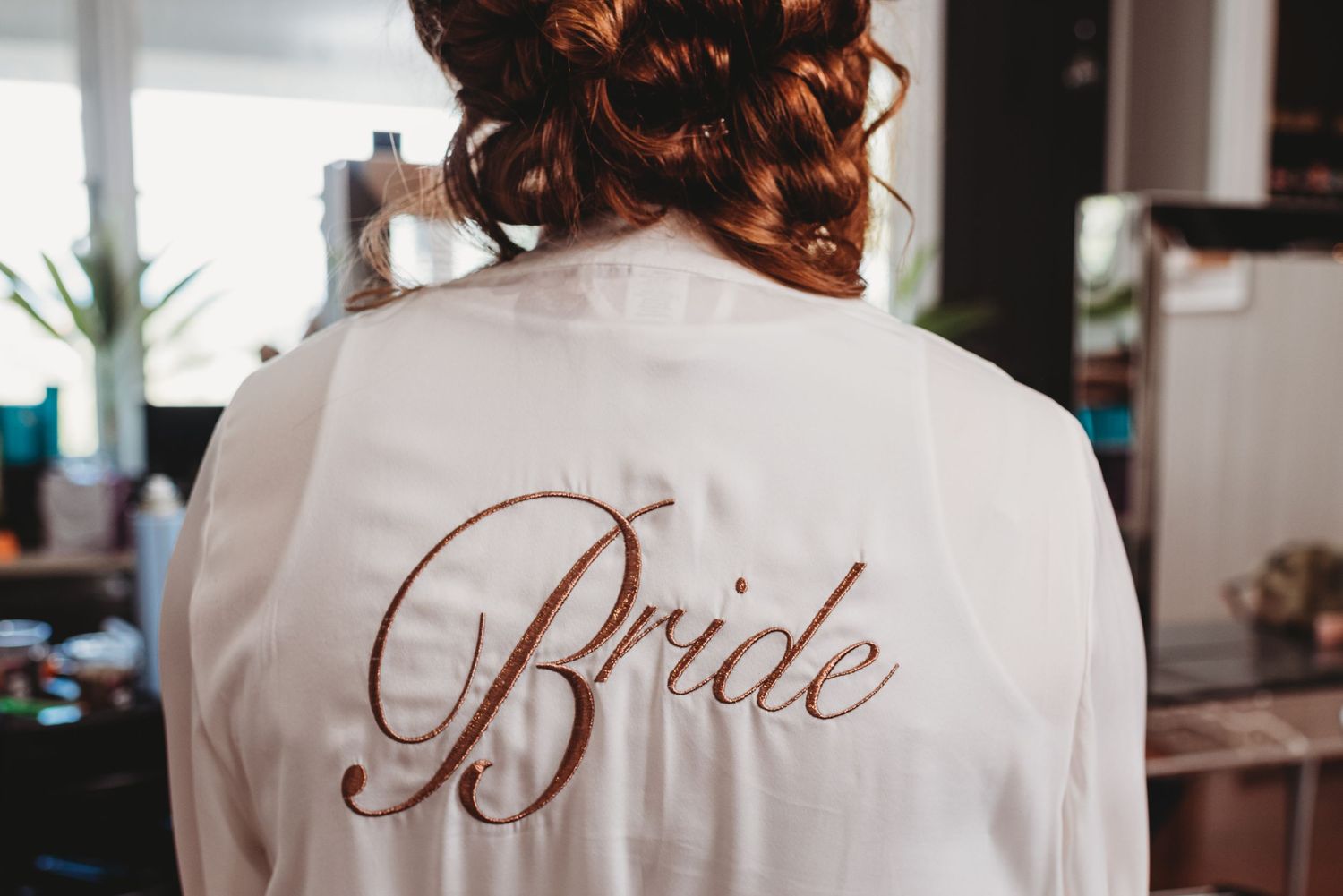 A white satin robe with 'Bride' embroidered in rose gold script.