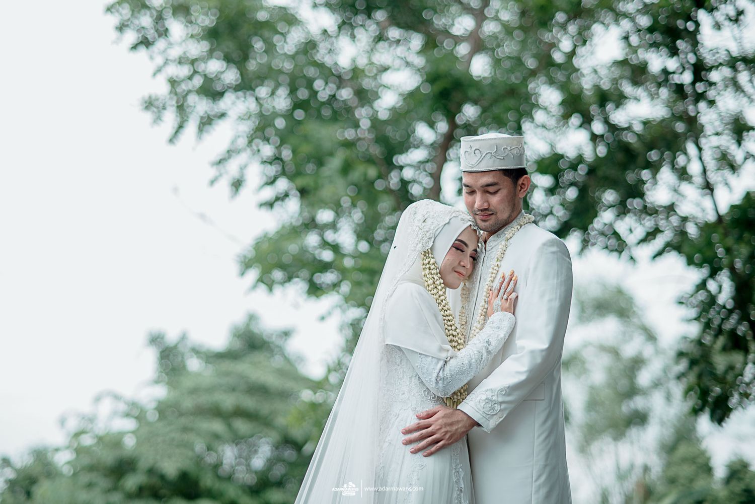 Couple in white Islamic wedding attire embracing outdoors with trees in background.