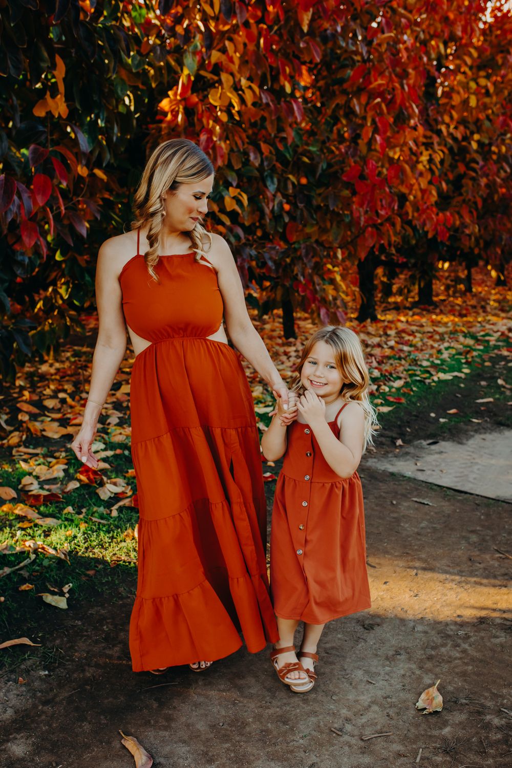 Mother and daughter in matching red dresses standing together among autumn trees.