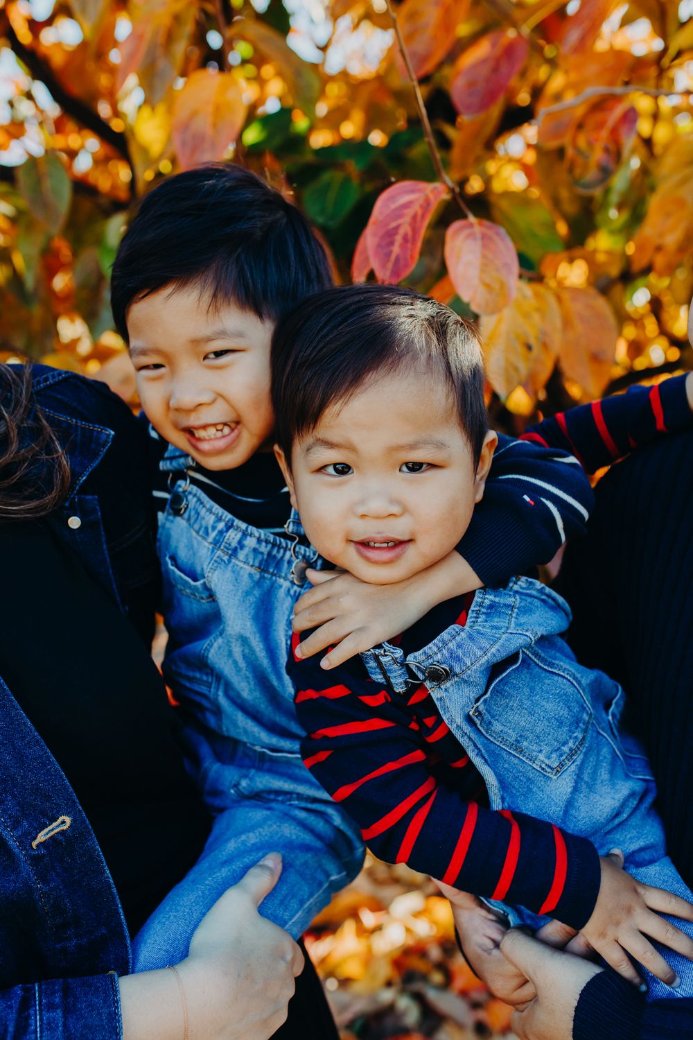 Two children in denim outfits smile brightly against a backdrop of autumn foliage.