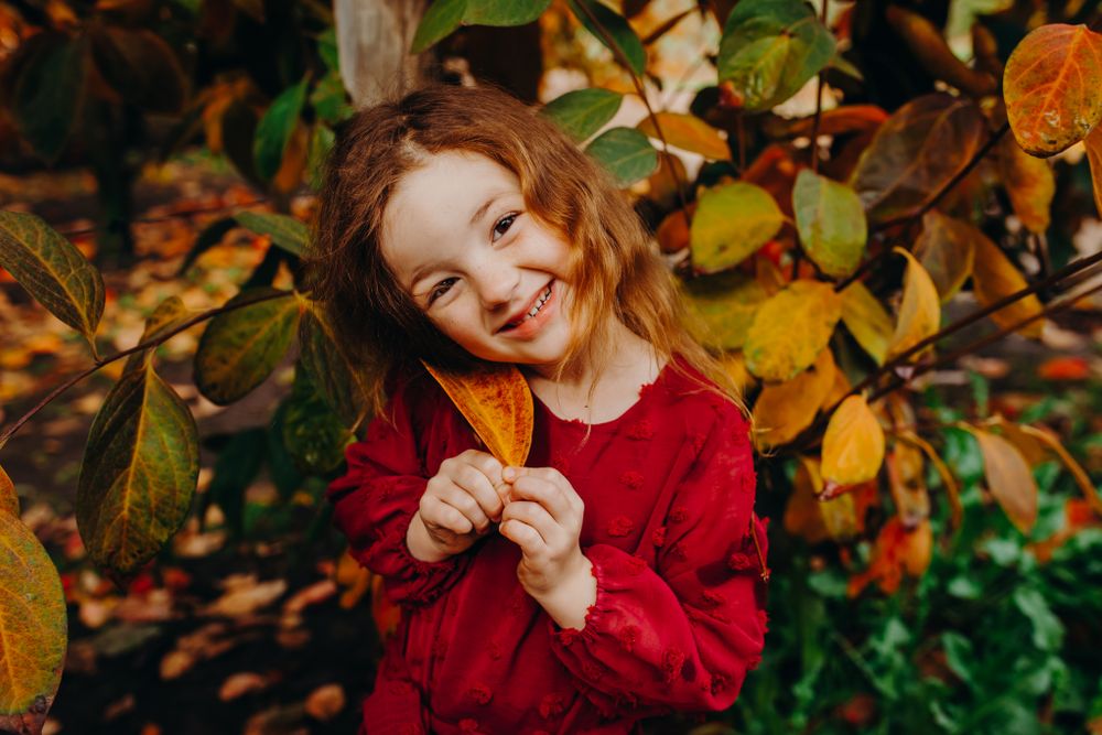 Child in red sweater smiling while holding autumn leaves against colorful fall background.
