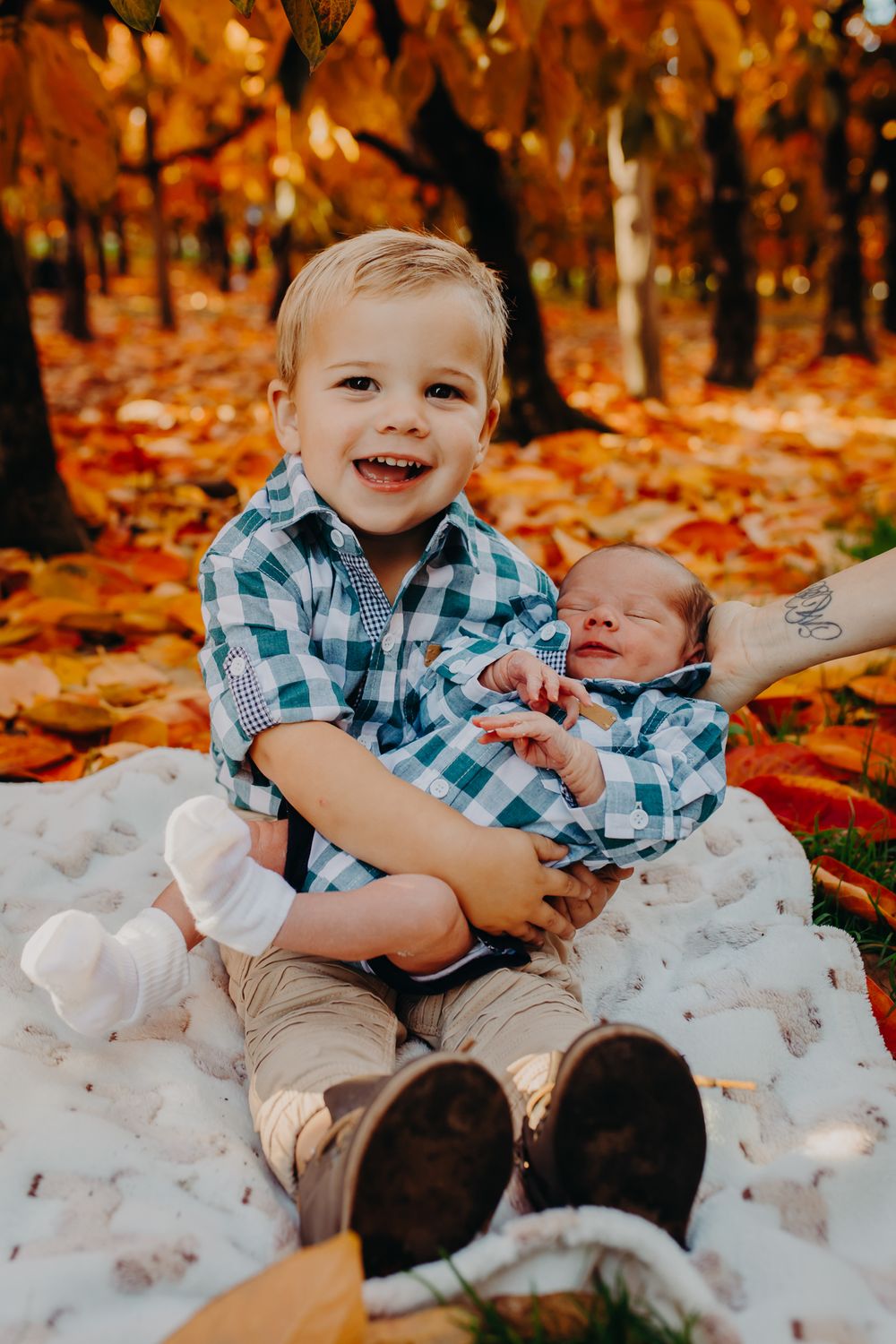 Young child holding infant sibling while sitting on blanket surrounded by orange fall leaves.