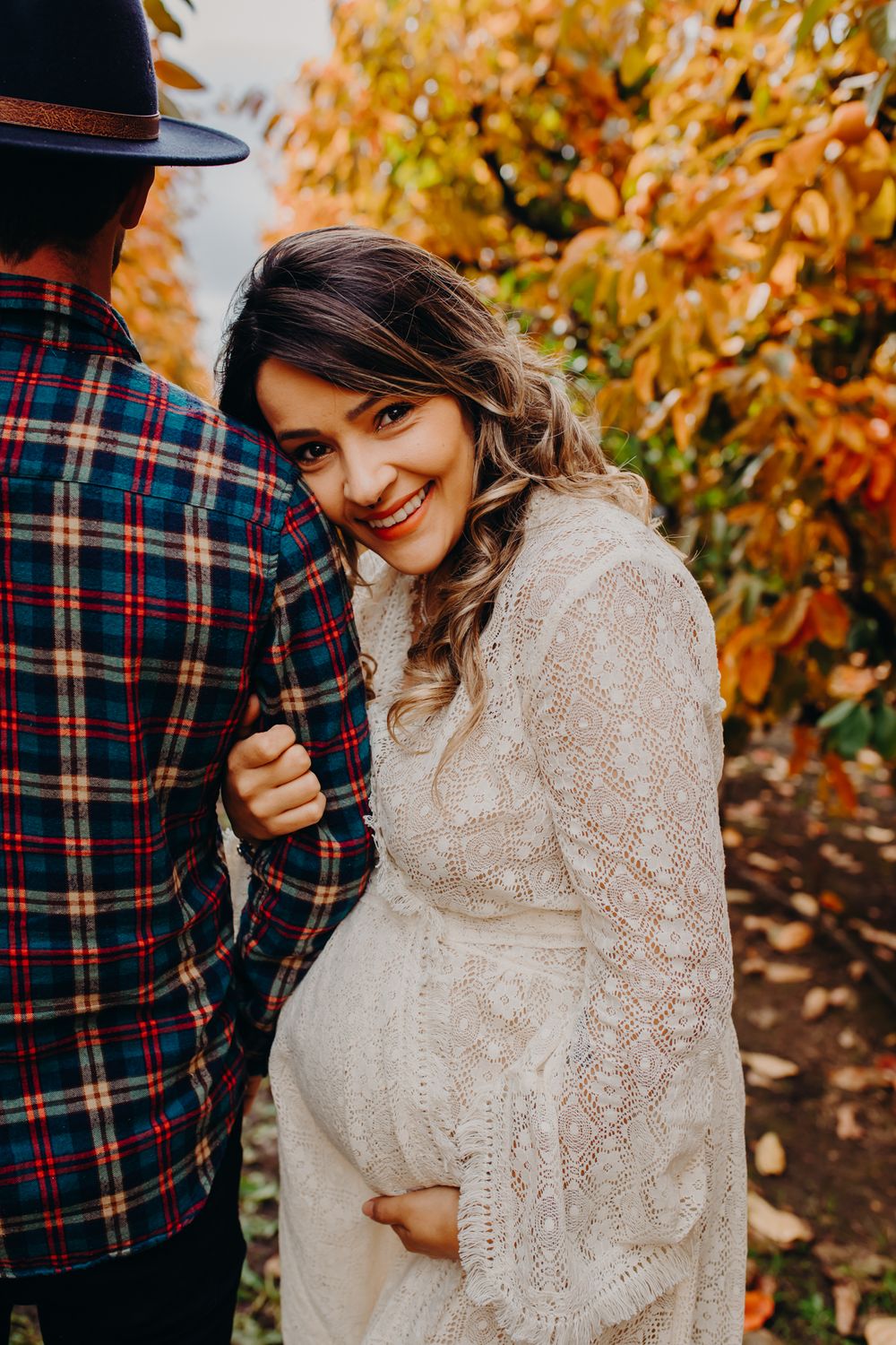 Expectant couple in casual attire poses among fall foliage.