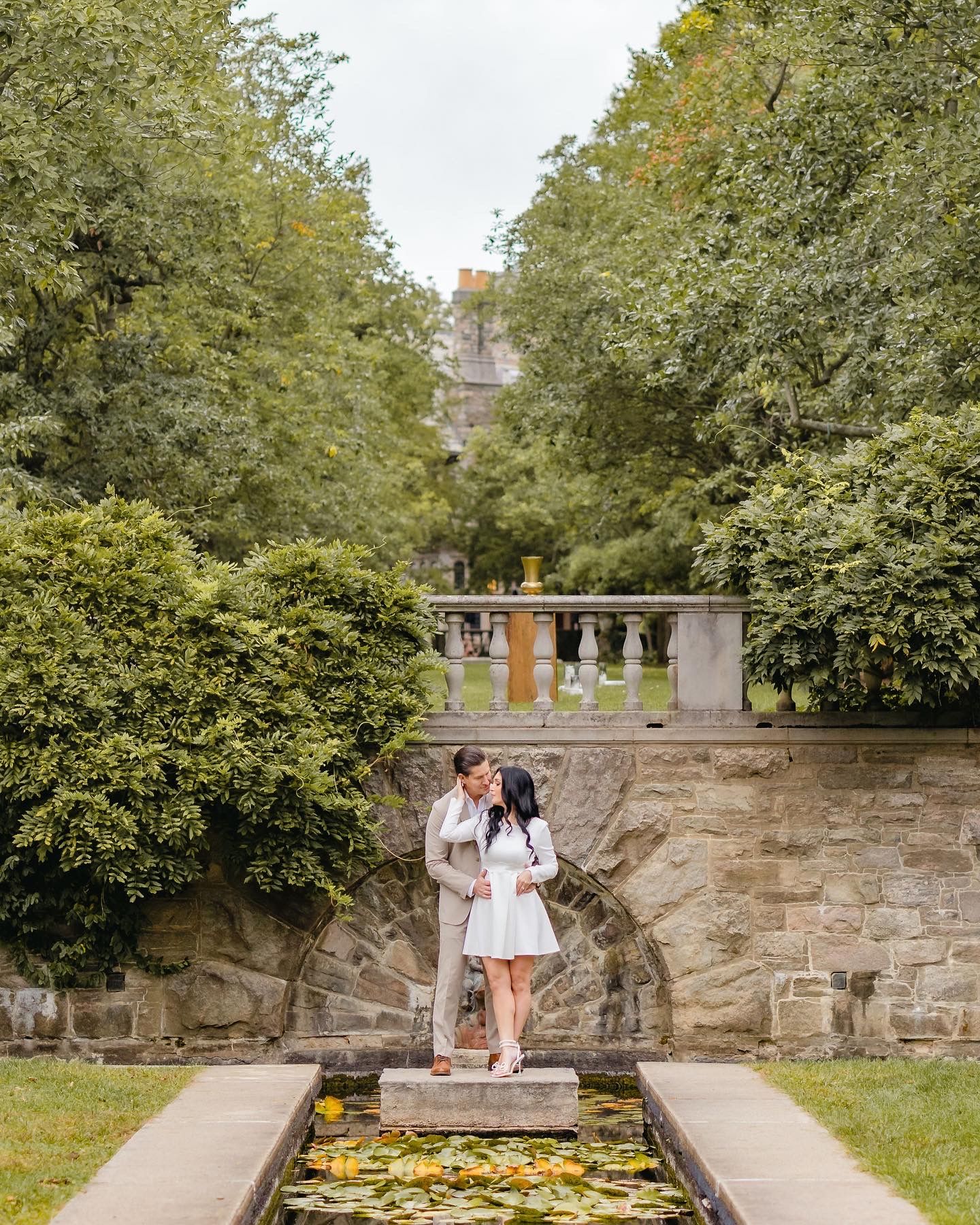 Couple sharing a kiss beside a lily pond in a lush garden with stone architecture.