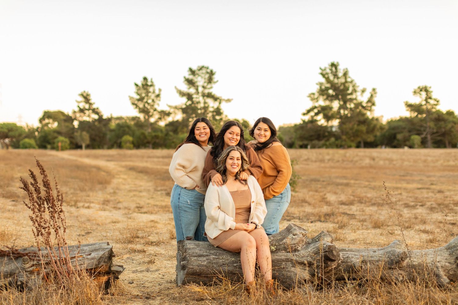 Friends gather together on a fallen log in a golden field during sunset.