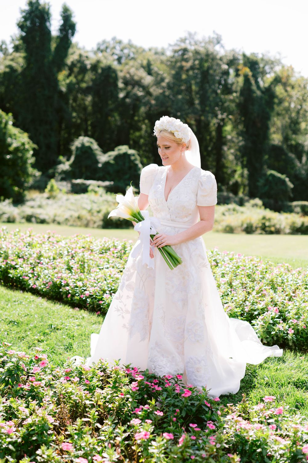 A bride stands among blooming rose gardens holding a simple white bouquet.