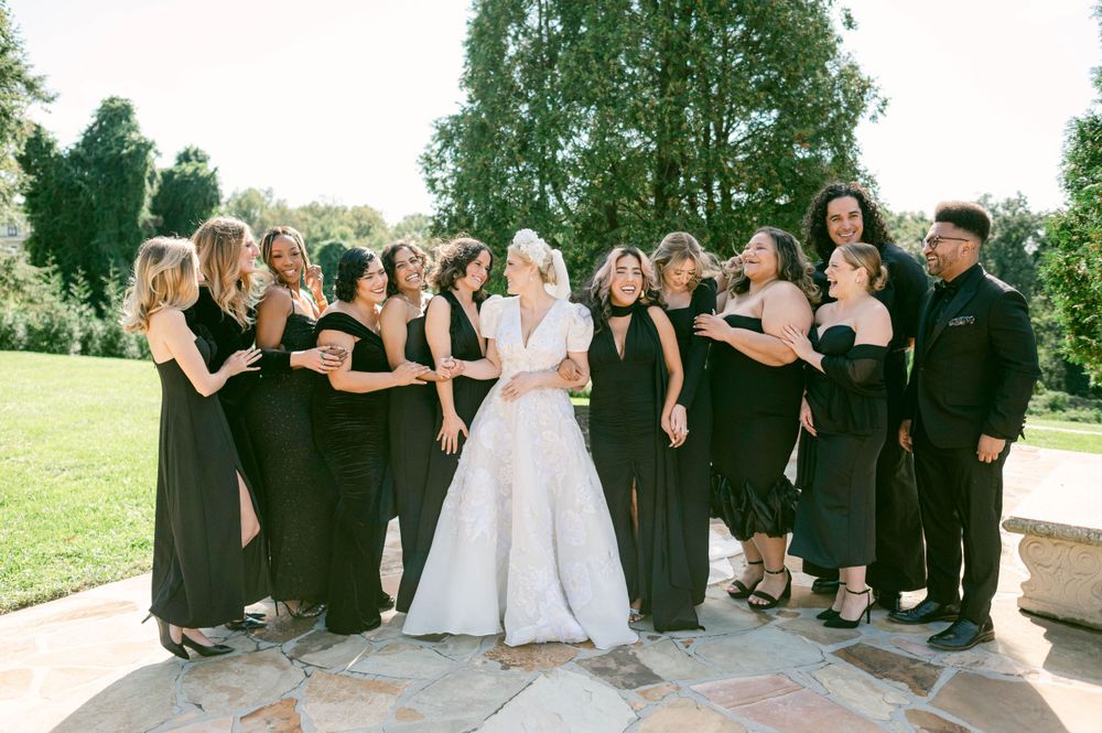 A wedding party in black attire poses with the bride in white on an outdoor stone patio at Oxon Hill Manor.